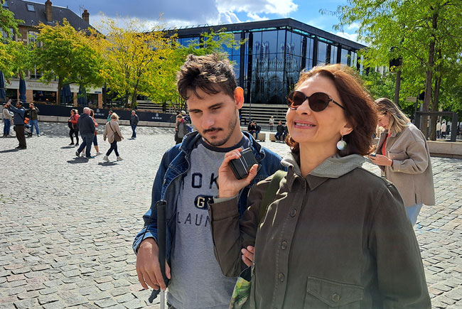 Sur un parvis ensoleillé, un femme au visage rayonnant porte des lunettes de soleil. Près d'elle, un jeune homme aux yeux clos, attentif. Dans sa main droite, une canne blanche. De la gauche, il tient le bras de sa voisine. Entre eux, elle maintient un téléphone portable à la hauteur de leurs oreilles. Derrière eux, sur la façade de verre d'un bâtiment, le reflet de la cathédrale Notre-Dame de Reims.