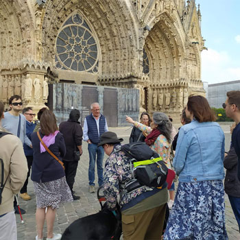 Sur le parvis de la cathédrale Notre-Dame de Reims, un peu groupe entoure une femme aux longs cheveux gris, veste fleurie. Elle pointe du doigt un détail de la façade principale. Deux visiteurs tendent le cou, une se penche vers son chien-guide. Un troisième, une canne blanche à la main, a le visage attentif.