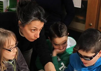 Trois enfants d'une dizaine d'années, assis en arc de cercle, attentifs. L'un d'entre eux porte des lunettes noires. Penchée au-dessus d'eux, Marie s'adresse à eux.